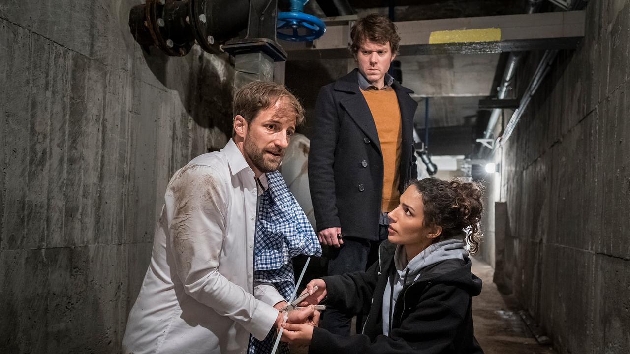 In einem Beton-Korridor kniet Philipp Baumgarten, während Kim Nowak ihm an den gefesselten Händen hilft. Tom Kowalski steht daneben.