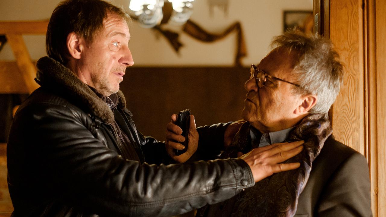 In einem rustikalen Raum packt Kurt Lehrmann in Lederjacke Dr. Johannes Mayerhofer am Kragen, während Mayerhofer einen Elektroschocker in der Hand hält.