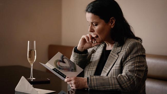 Ende 50-jährige Frau, sitzt an einem Tisch, vor ihr ein Glas Sekt, sie hält nachdenklich eine Karte in der Hand