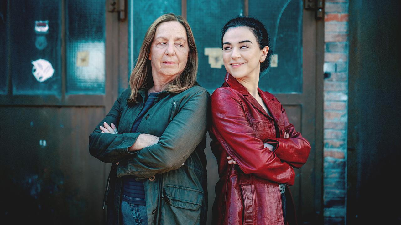 Gerti Bruckner und Heidi Mai stehen Rücken an Rücken vor einer industriellen Tür, die Arme verschränkt, in einem kühl-bläulichen Licht.