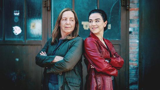 Gerti Bruckner und Heidi Mai stehen Rücken an Rücken vor einer industriellen Tür, die Arme verschränkt, in einem kühl-bläulichen Licht.