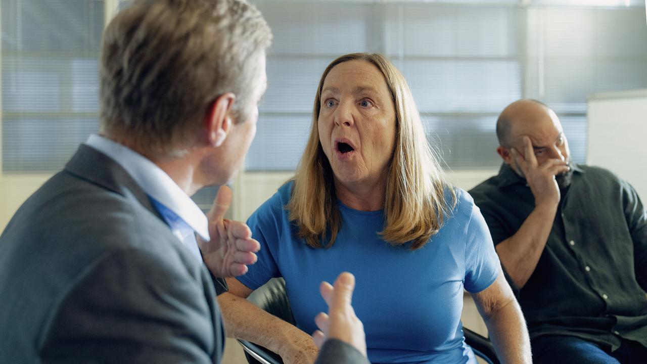 In einem Seminarraum Gerti mit jemanden im Vordergurnd, während Adam Reza neben Gerti sitzt.