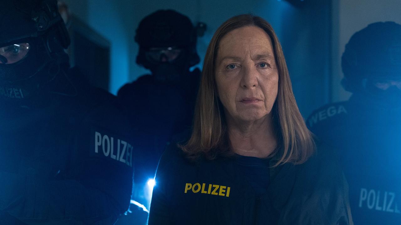 In blauem Licht steht Gerti in Polizeiuniform, hinter ihr sind schwer ausgerüstete Einsatzkräfte zu sehen.