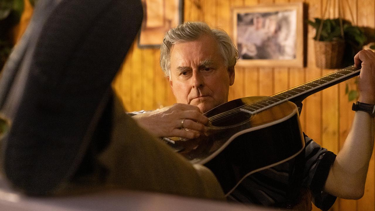 Bertl  sitzt mit einer akustischen Gitarre in der Hand in einem warm beleuchteten Raum vor einer Holzpanelwand.