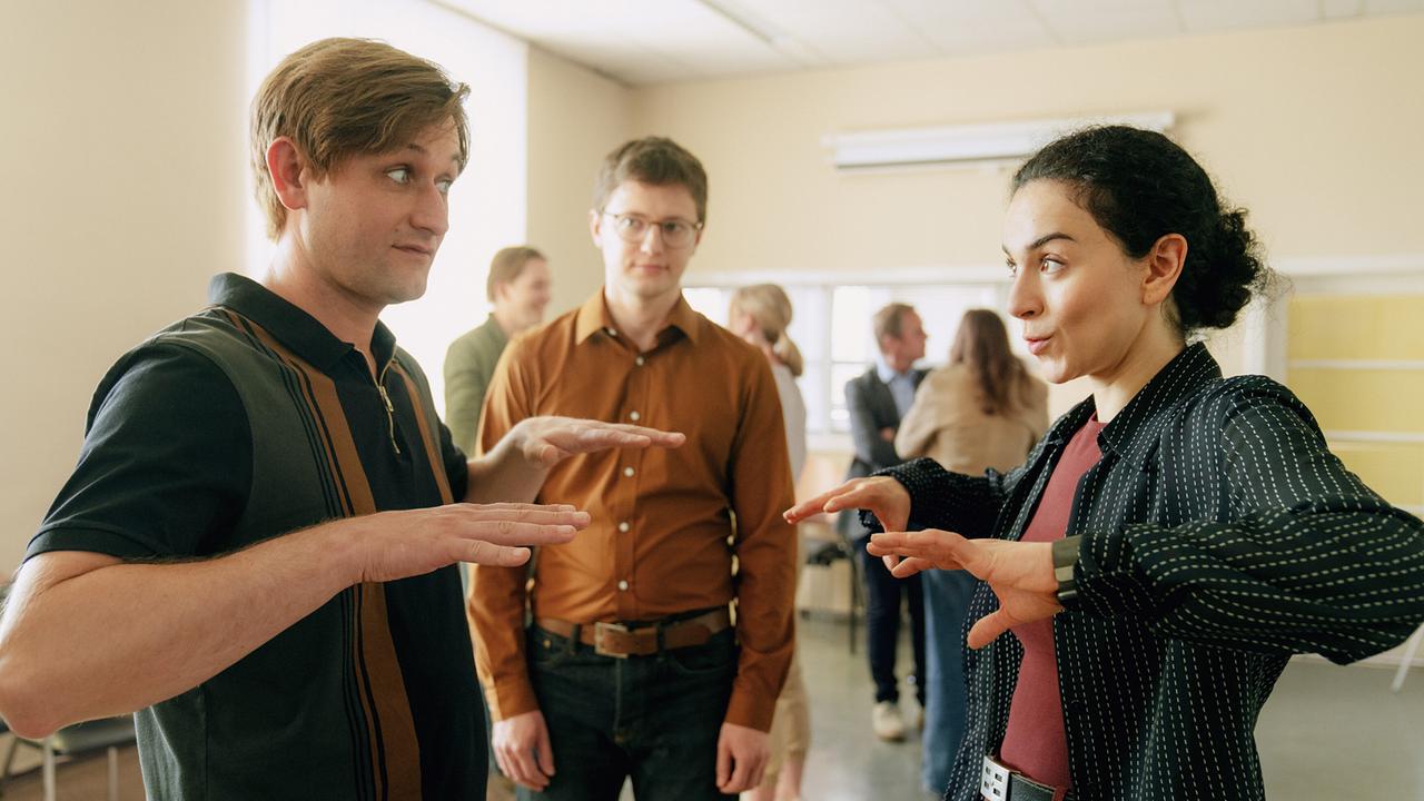 Harald Vogel und Heidi stehen sich im Seminarraum mit angewinkelten Armen gegenüber, während Coach Rainer ihnen zusieht und andere im Hintergrund zu sehen sind.