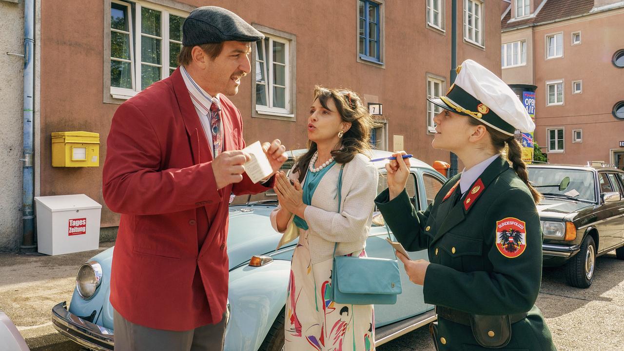 Auf einer Straße spricht eine uniformierte Gerti in den 80ern mit einem Parksünder und deren Gattin neben einem hellblauen VW-Käfer.