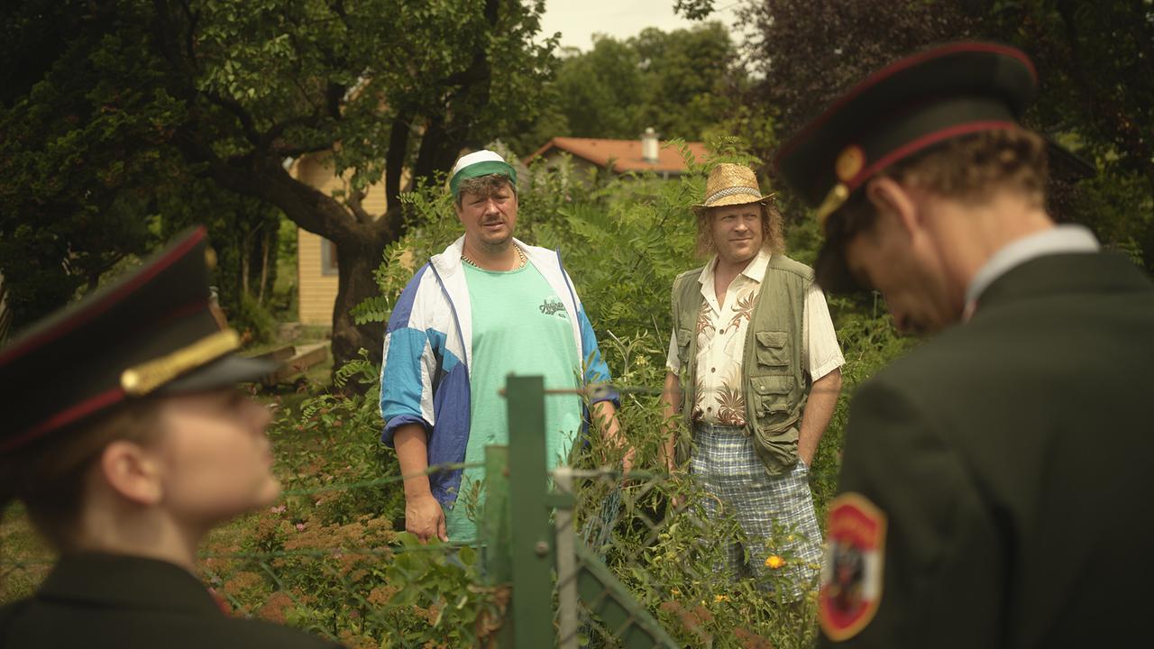Gerti und Kranzler, beide in Polizeiuniform, stehen in den 80er einem Garten am Zaun, an dem dahinter zwei Männer zwischen dichtem Grün in einem Kleingarten stehen.