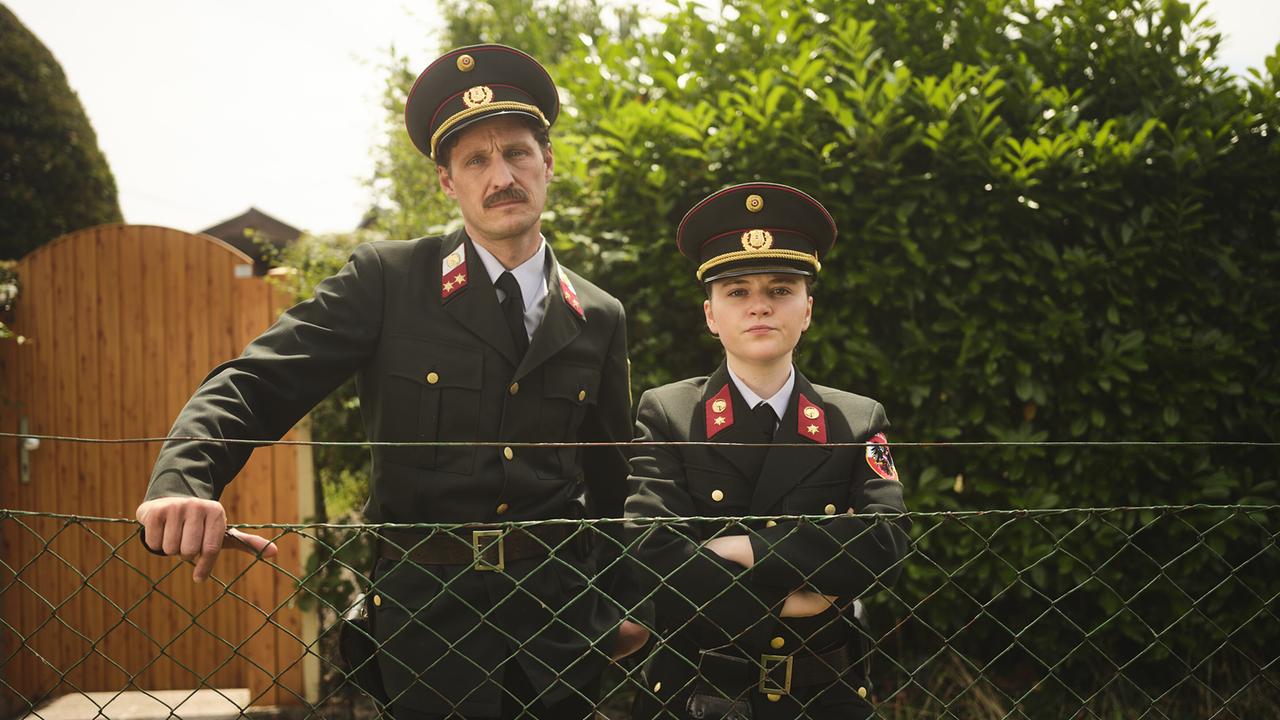 Kranzler und Gerti, beide in Polizeiuniform, stehen in den 80ern an einem Maschendrahtzaun vor dichtem Gebüsch in einer Kleingartensiedlung.