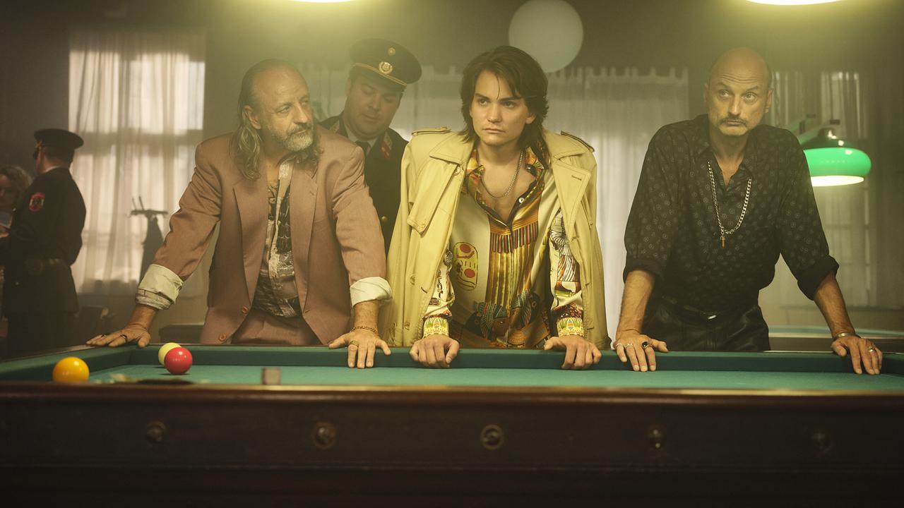 Drei Strizzis lehnen in den 80ern einer rauchigen Billardhalle an einem Billardtisch unter grünen Lampen, dahinter sind Polizisten zu sehen.