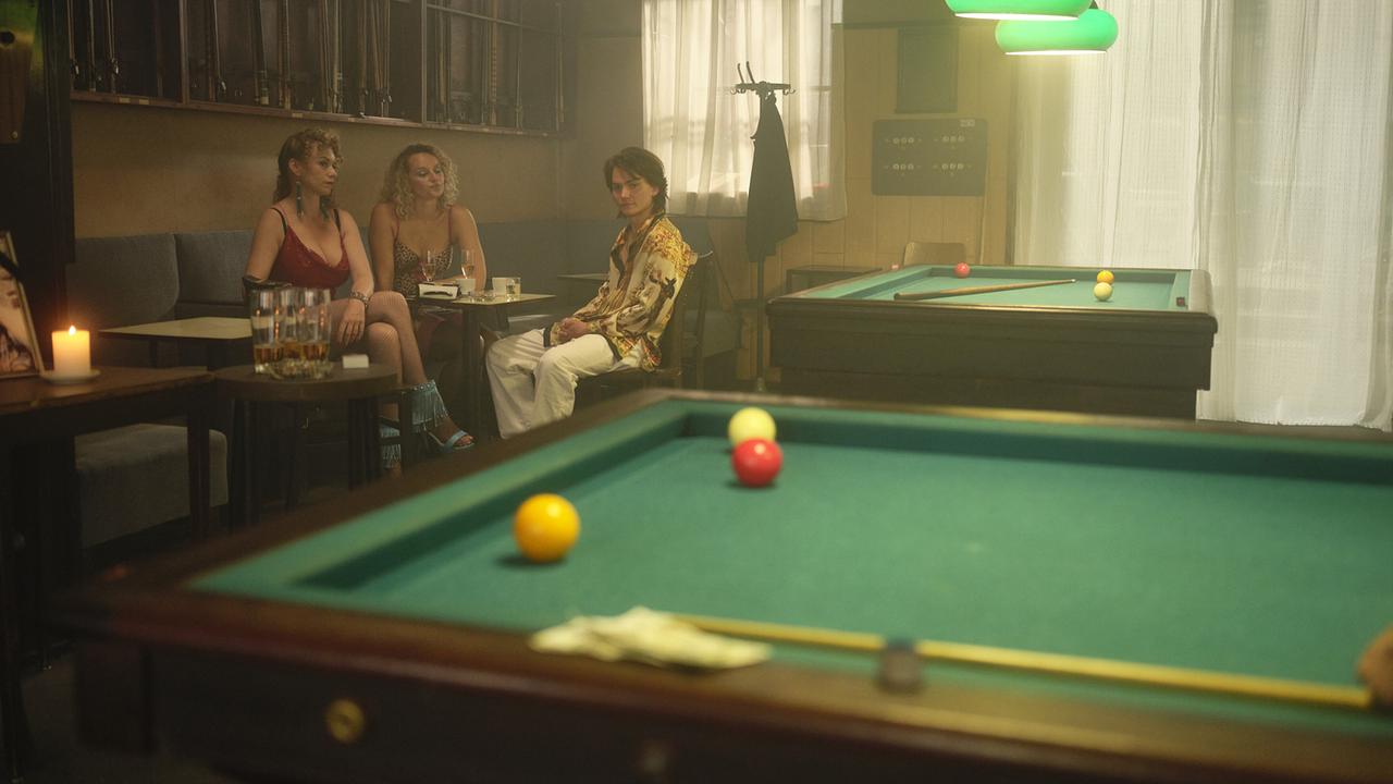 Lolo, Stella und Fanco sitzen in den 80ern in einem Billard-Café an einer Sitzbank, im Vordergrund liegt ein Billardtisch mit Kugeln.