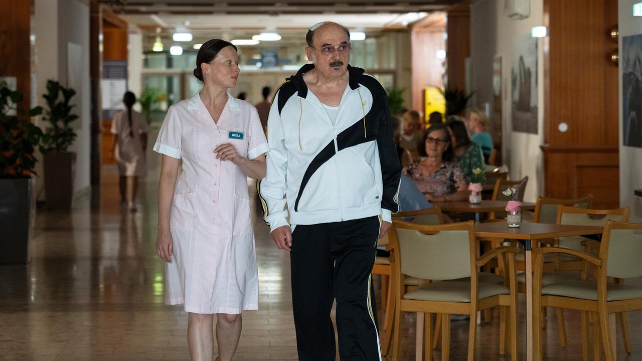 In einem Seniorenheim geht eine Pflegerin neben Franco im Trainingsanzug einen Flur entlang, im Hintergrund sitzen weitere Personen.