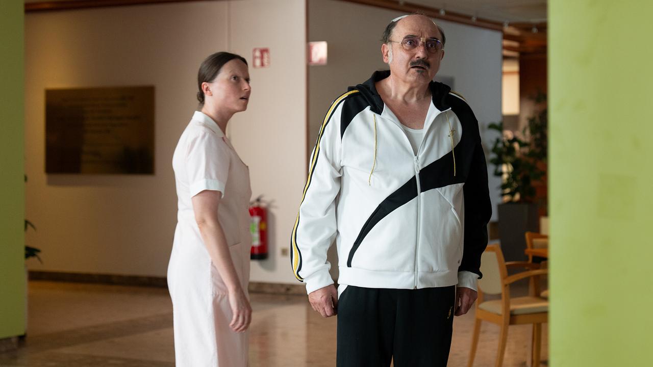 Franco im Trainingsanzug steht in einem Flur im Seniorenheim, während eine Pflegerin ihn von hinten anschaut.