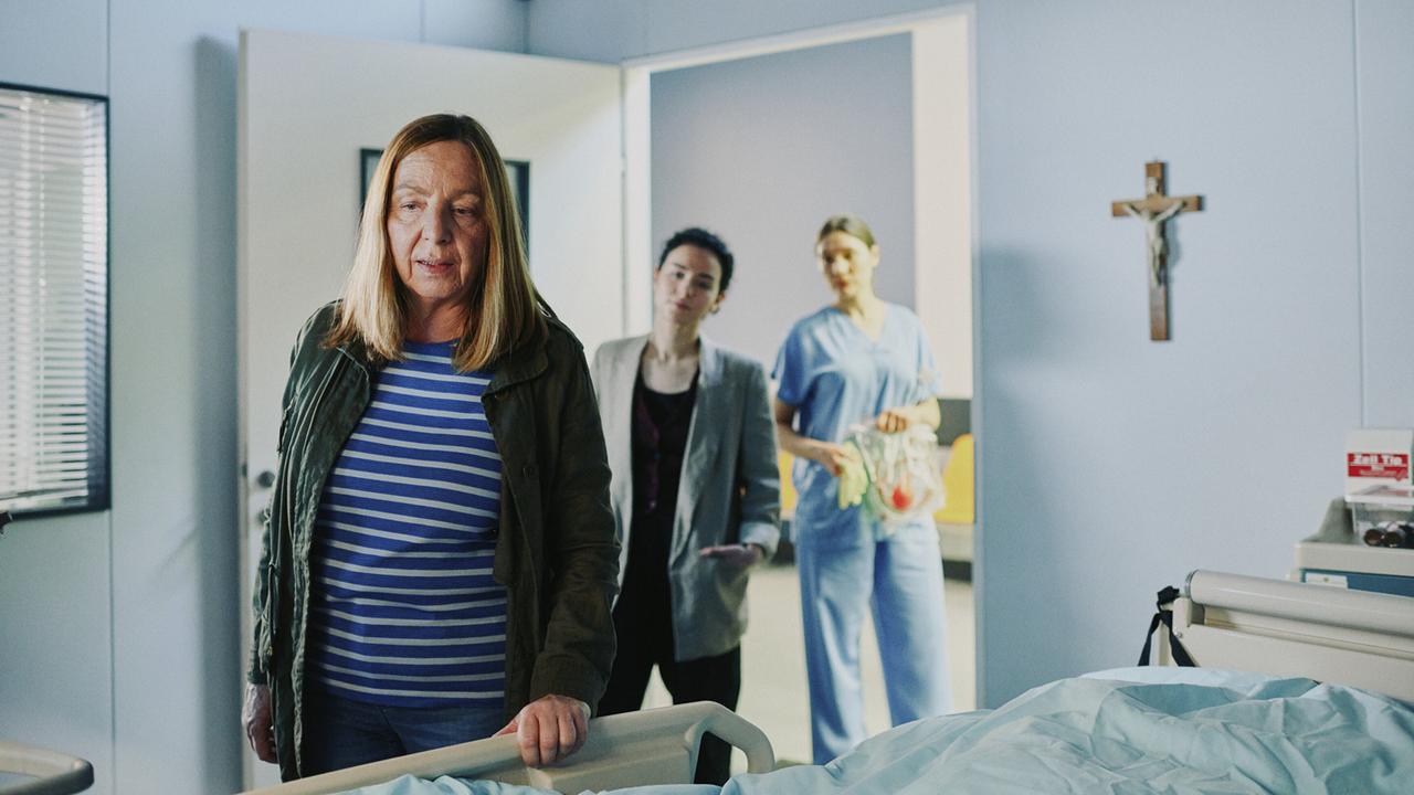 In einem Krankenzimmer mit einem Kruzifix an der Wand steht Gerti am Bett, in der Tür stehen Heidi und eine Krankenschwester.
