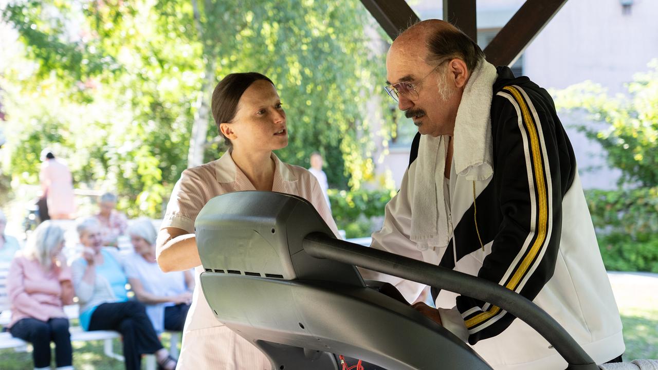Draußen trainiert Franco auf einem Laufband bei Sonnenschein, während eine Pflegekraft ihn betreut und andere Bewohner:innen im Hintergrund sitzen.