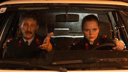 Durch die Windschutzscheibe eines Polizeiautos sind Gerti am Lenkrad und Kranzler am Beifahrersitz, der gestikuliert spricht, zu sehen.