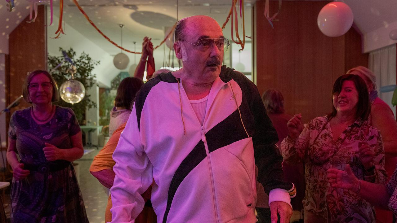 Franco betritt einen Partyraum, in dem mehrere Frauen in einem festlich geschmückten Raum mit Luftballons, Girlanden und farbigem Partylicht tanzen.