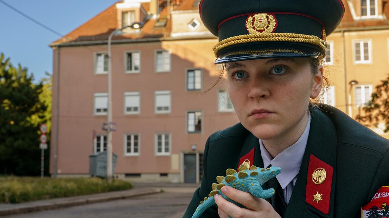 Die uniformierte junge Gerti hält in den 80ern eine kleine Spielzeug-Echse in der Hand, vor einer Straße mit Wohngebäuden im Hintergrund.