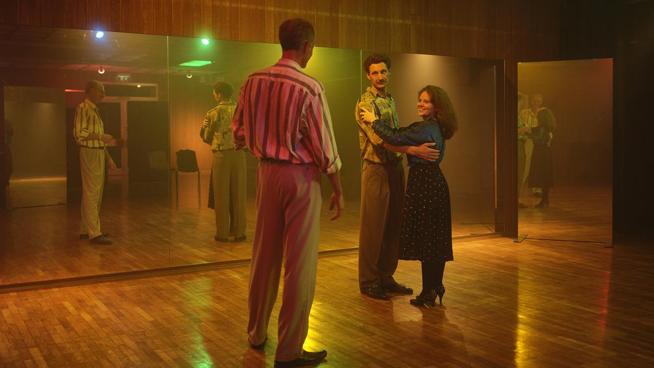 In einem warm beleuchteten Tanzsaal mit einer Spiegelfront stehen in den 80ern Kranzler und Gerti in Paarhaltung beieinander. Vor ihnen steht ein Tanzlehrer.