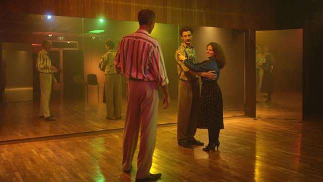 In einem warm beleuchteten Tanzsaal mit einer Spiegelfront stehen in den 80ern Kranzler und Gerti in Paarhaltung beieinander. Vor ihnen steht ein Tanzlehrer.