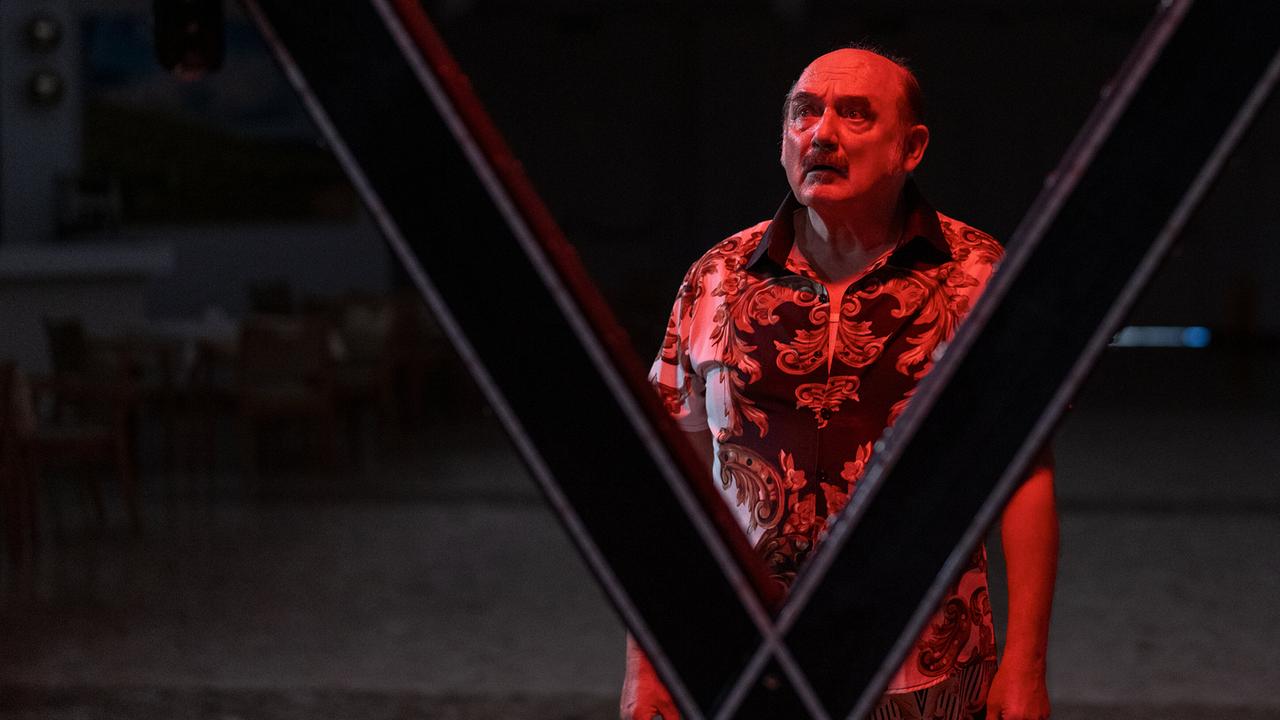 Franco in auffälligem gemustertem Hemd steht nachts unter rotem Licht hinter einer gekreuzten Metallkonstruktion in einer dunklen Umgebung.