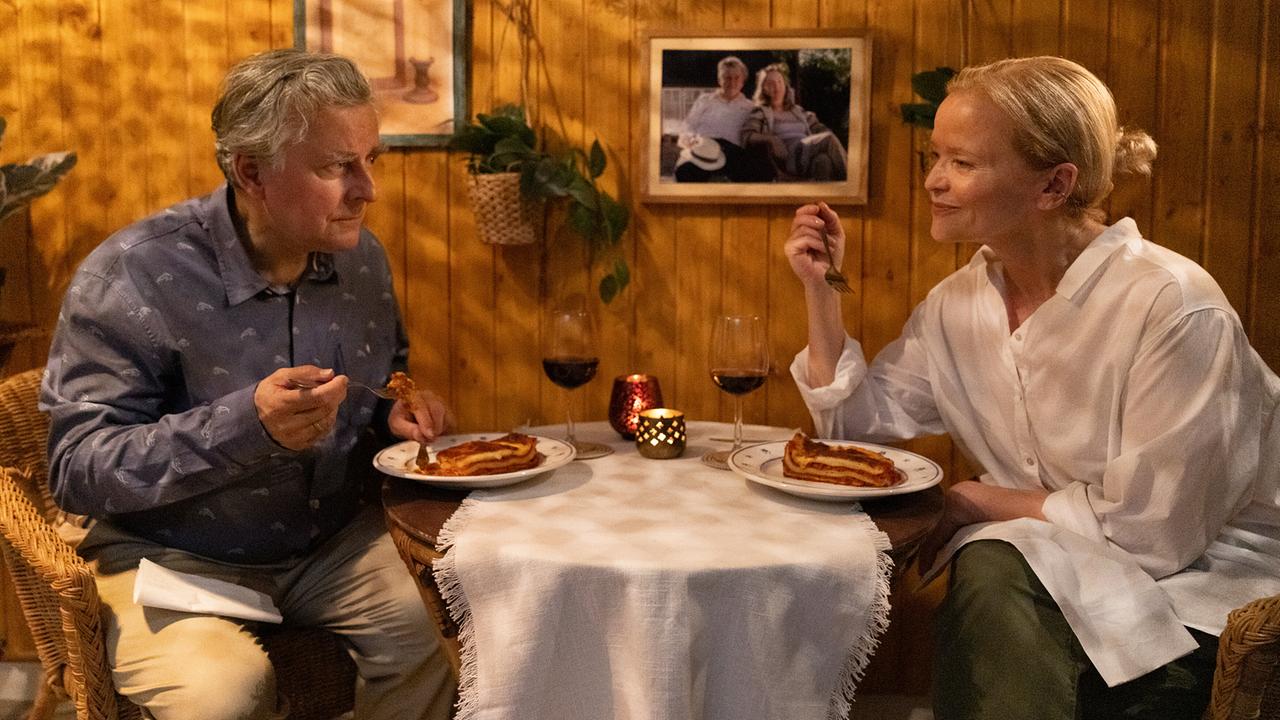 Bertl und Nora sitzen in Korbsesseln an einem kleinen Tisch in einem gemütlichen, holzvertäfelten Raum und essen gemeinsam bei Kerzenlicht und einem Glas Rotwein.