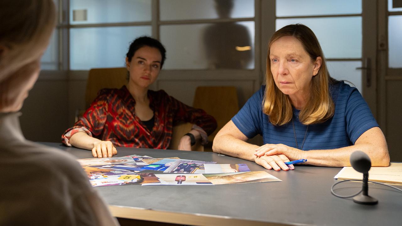 Heidi und Gerti sitzen in einem geschlossenen Raum an einem Tisch mit einer weiteren Frau, die unscharf im Vordergrund zu sehen ist. Auf dem Tisch liegen Beweisfotos soweie ein Aufnahmegerät mit Mikrofon.