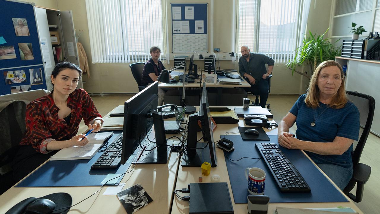 Innenansicht des Ermittlungsbüros und vier Schreibtischen mit Computern, an denen sich an den zwei vorderen Schreibtischen Heidi und Gerti gegenübersitzen und an den zwei hinteren Harald Vogel und Adam Reza.