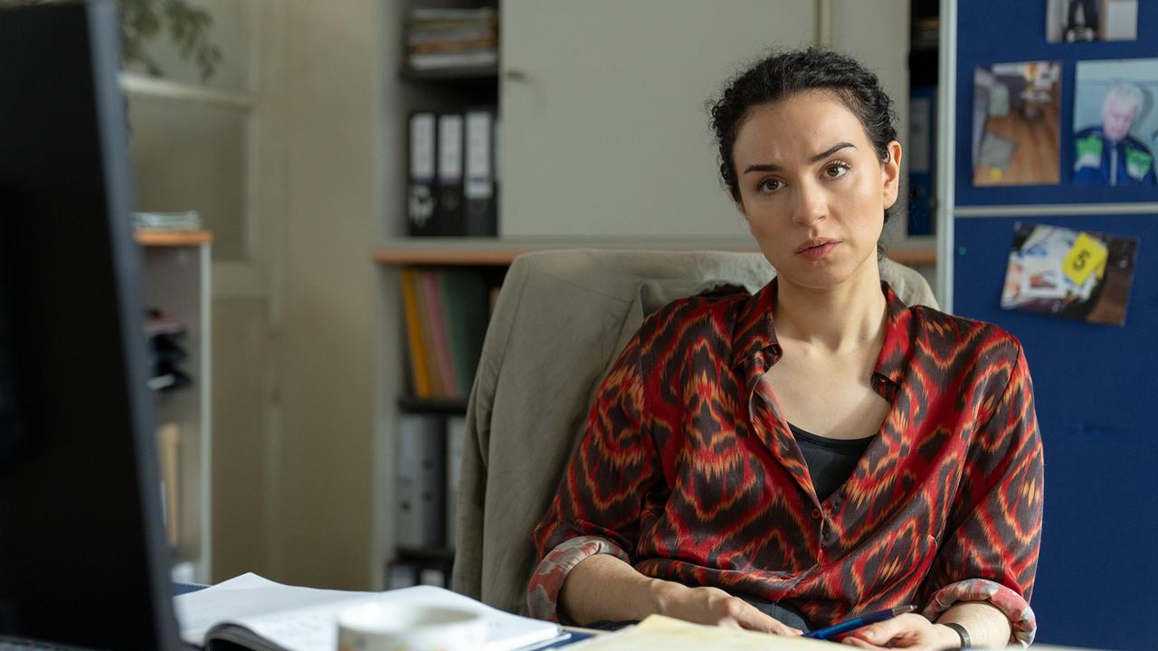 Heidi sitzt an ihrem Schreibtisch mit Unterlagen und Stift in einem Arbeitszimmer voller Ordner und Fotos an einer Pinnwand.