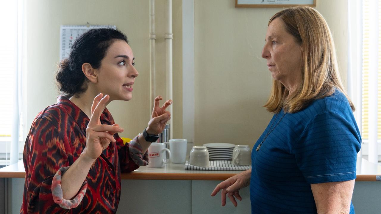 Heidi und Gerti unterhalten sich gestikulierend vor einer Anrichte mit Tassen und Geschirr in einem hellen Raum.