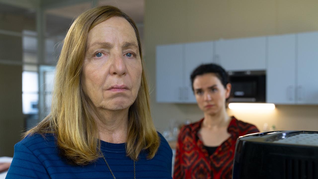 In einer Büroküche stehen Gerti im Vordergrund und Heidi dahinter beisammen, während im Vordergrund eine Kaffeemaschine auf der Arbeitsfläche zu sehen ist.