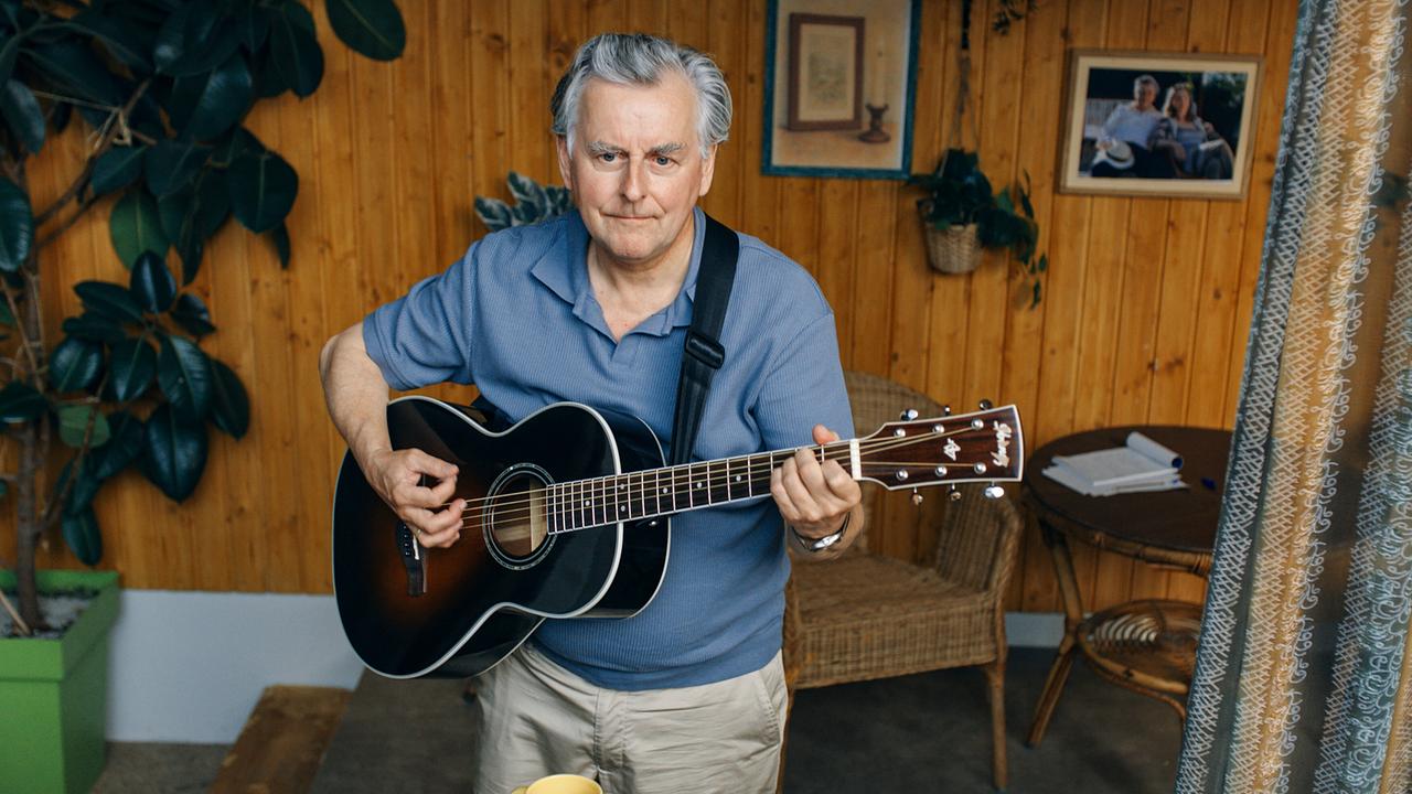 Bertl spielt in einem holzvertäfelten Raum auf einer Akustikgitarre, umgeben von Pflanzen, Möbeln und persönlichen Gegenständen.