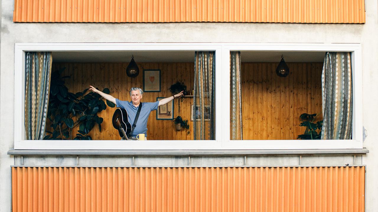 Bertl  steht in einem offenen Fenster seiner Wohnung mit ausgebreiteten Armen und einer Gitarre umgehängt.