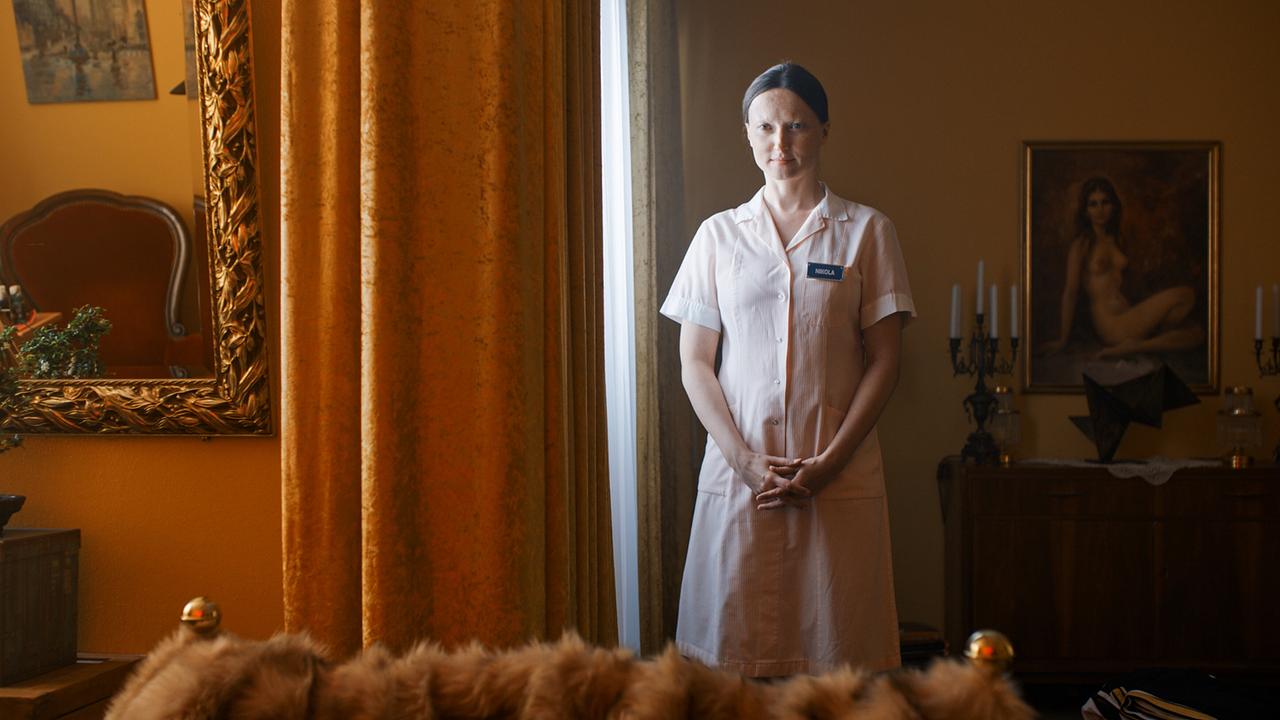 Die Pflegerin Nikolat steht mit gefalteten Händen in einem stilvoll eingerichteten Zimmer mit schweren Vorhängen, Kerzenleuchtern und klassischer Dekoration.