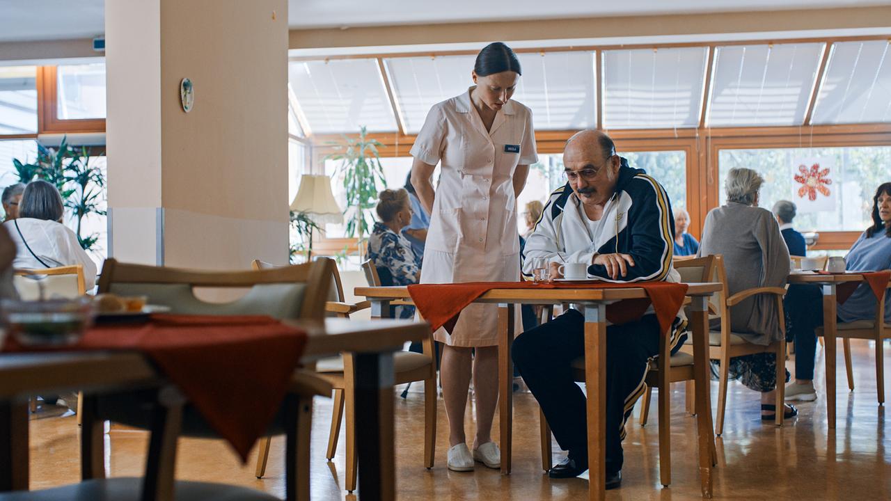 In einem Speisesaal einer Betreuungseinrichtung sitzt Franco an einem Tisch, während Pflegerin Nikolat neben ihm steht und andere Bewohner:innen im Hintergrund zu sehen sind.