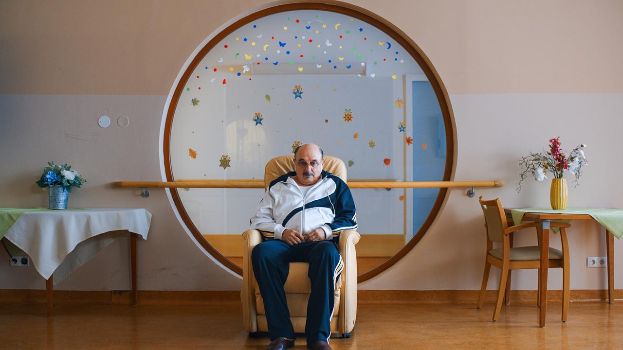 Franco sitzt in einem Ledersessel vor einem großen, runden Sichtfenster zwischen Tischen und Dekoration in einem hellen Aufenthaltsraum.