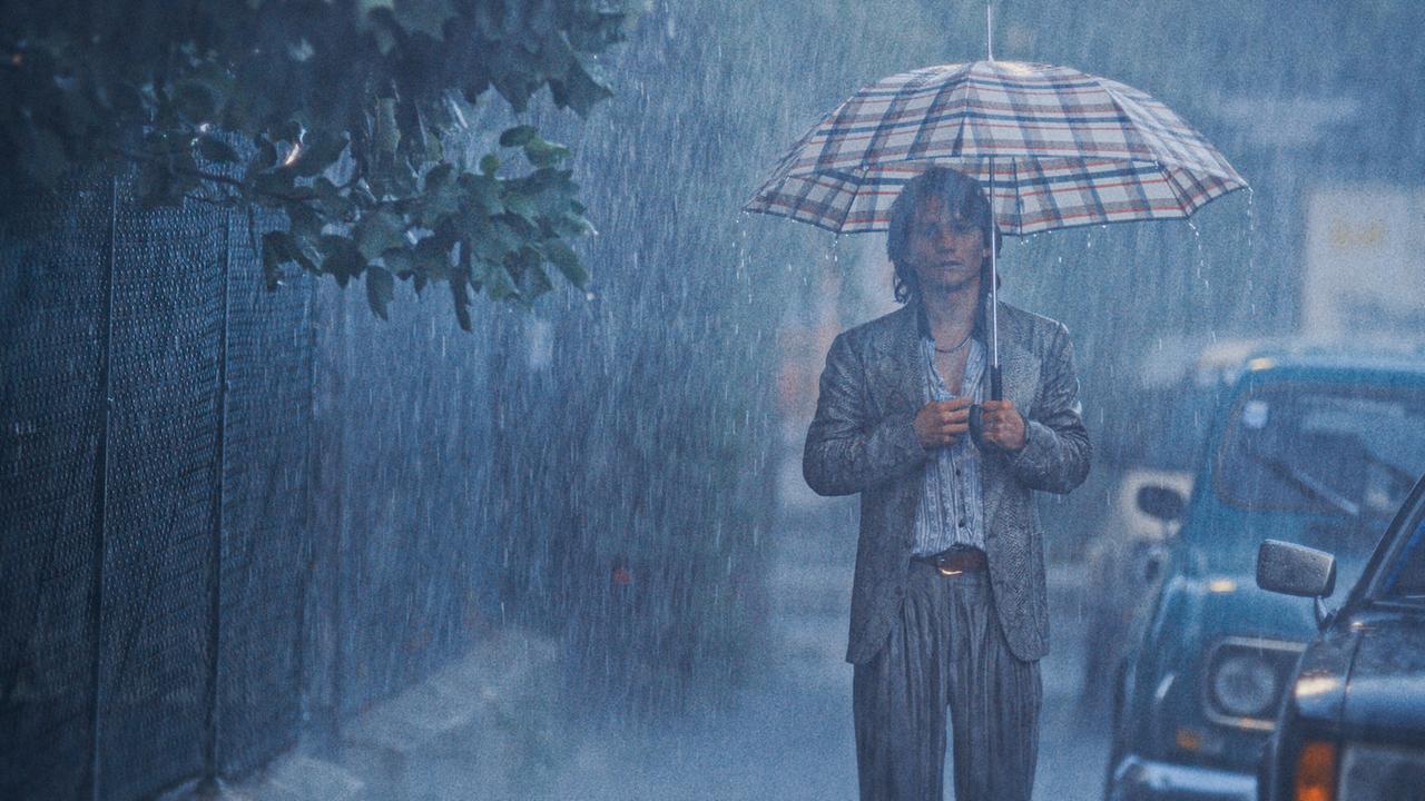 Franco steht in den 80ern bei starkem Regen mit einem Regenschirm in einer ruhigen Straße, während parkende Autos im Hintergrund zu sehen sind.