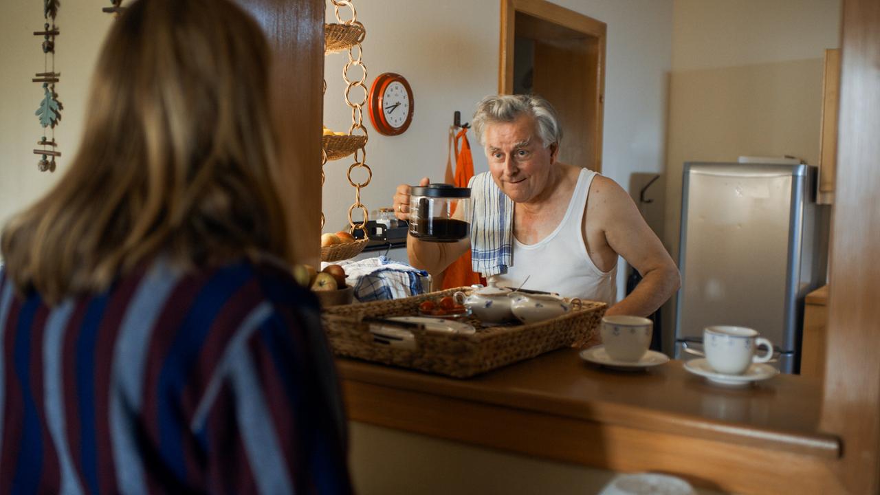 © ORF/Lotus Film In einer Küche steht Bertl hinter der Theke und hält eine Kaffeekanne, während Gerti ihm auf der anderen Seite der Theke gegenübersteht.