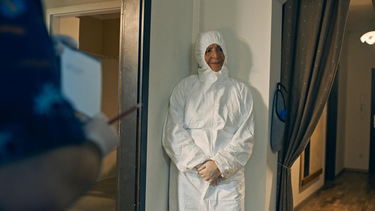 © ORF/Lotus Film In einer Ecke steht Gerti in Schutzkleidung an der Wand, während im Vordergrund jemand mit Handschuhen etwas notiert.