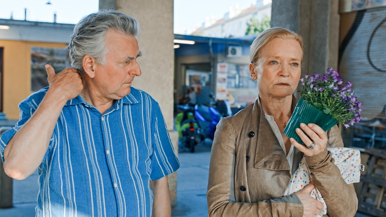 © ORF/Lotus Film Bertl steht neben Nora, die einen kleinen Blumentopf hält, im Freien vor einem Gebäude.