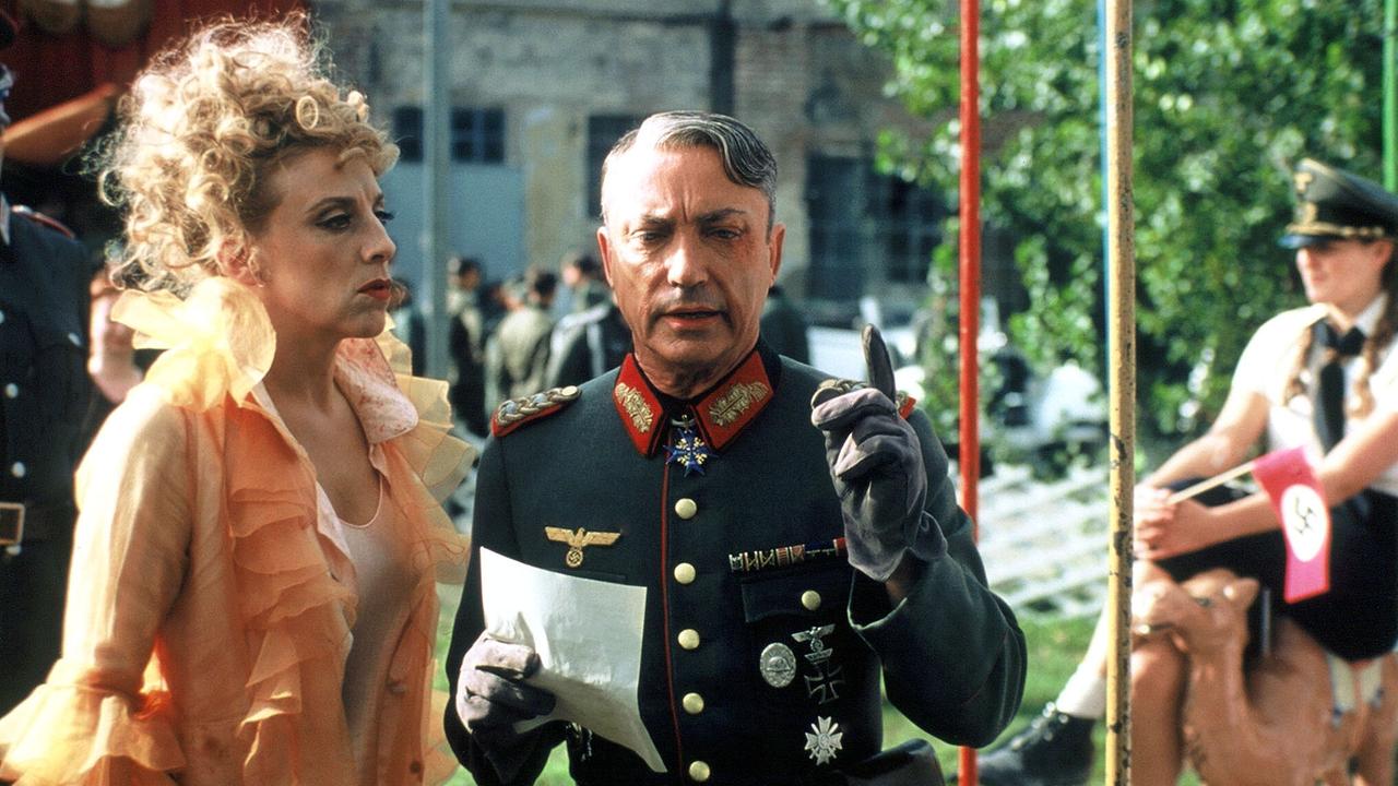 Sissi Perlinger in einer auffälligen, rüschenbesetzten Kleidung steht im Freien neben Udo Kier in einer historischen Militäruniform aus der NS-Zeit, der ein Dokument hält und gestikuliert.