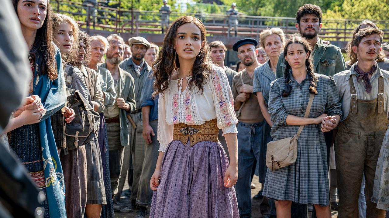© ORF/LEONINE Studios/© 2023 Lionsgate/Murray Close Eine junge Frau steht vor einer Gruppe von Menschen in einem von bewaffneten Personen bewachten Areal, alle schauen verunsichert und sorgenvoll in dieselbe Richtung