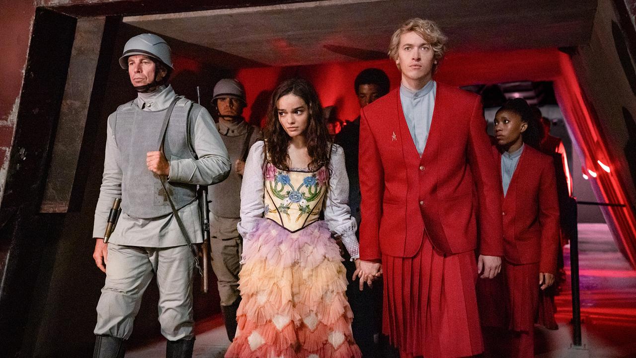 © ORF/LEONINE Studios/© 2023 Lionsgate/Murray Close Rachel Zegler als Lucy Gray Baird in einem pastellfarbenen bunten Kleid geht scheu an der Hand von Tom Blyth als Corolanus Snow in rotem Uniform-Anzug mit blaugrauem Hemd, begleitet von Soldaten und anderen uniformierten Personen