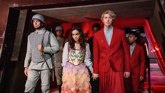 Rachel Zegler als Lucy Gray Baird in einem pastellfarbenen bunten Kleid geht scheu an der Hand von Tom Blyth als Corolanus Snow in rotem Uniform-Anzug mit blaugrauem Hemd, begleitet von Soldaten und anderen uniformierten Personen