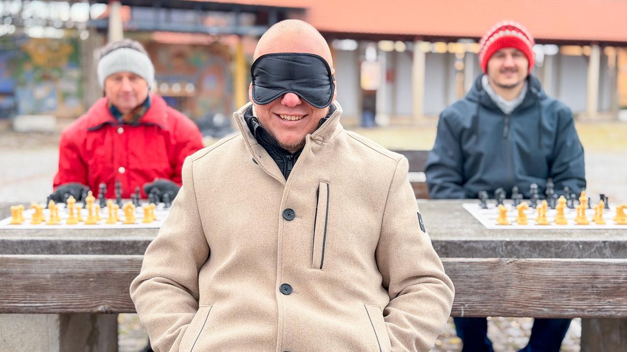 © ORF/BFILM Der österreichische Schach-Großmeister Markus Ragger mit verbundenen Augen sitzt an einem Tisch zwei Schachbrettern im Freien. Im Hintergrund sitzen zwei Männer am Tisch.