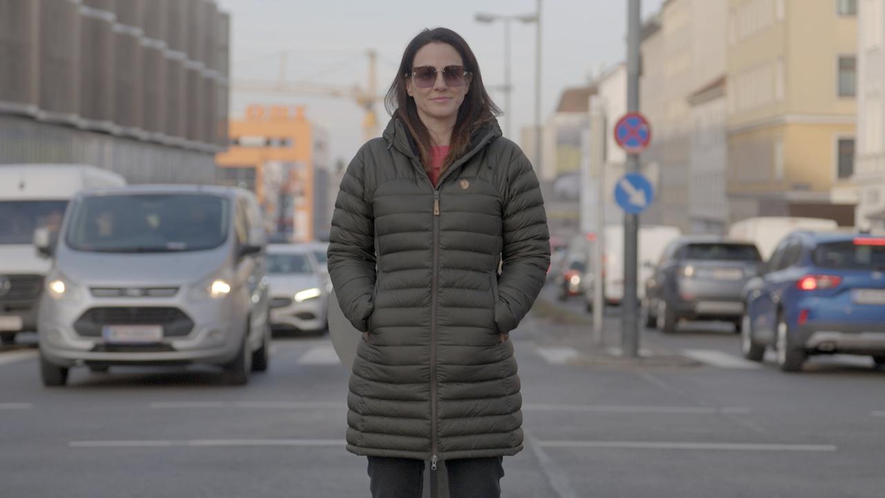 © ORF/BFILM Lisa Gadenstätter steht mit den Händen in den Taschen einer Winterjacke auf einer belebten Stadtstraße, während Autos und Gebäude den Hintergrund bilden.