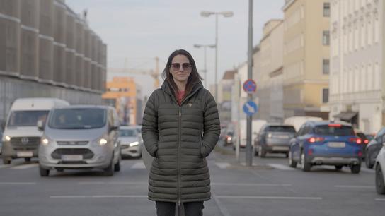 Lisa Gadenstätter steht mit den Händen in den Taschen einer Winterjacke auf einer belebten Stadtstraße, während Autos und Gebäude den Hintergrund bilden.