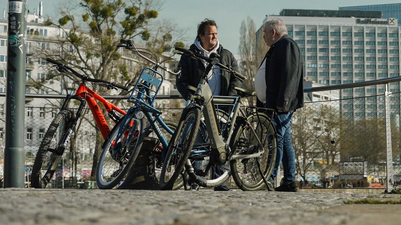 © ORF/HolyScreen Media/Stefan Domnanovich Hanno Settele und Privatdetektiv Gerrit Walgemoet stehen mit drei Lockvogel-Rädern in einer städtischen Umgebung.