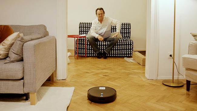 Hanno Settele sitzt in einer hellen Wohnung auf einem Sofa, während ein Saugroboter vor ihm über den Parkettboden fährt.