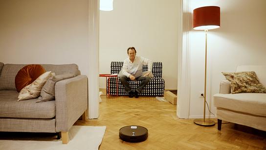 Hanno Settele sitzt in einer hellen Wohnung auf einem Sofa, während ein Saugroboter vor ihm über den Parkettboden fährt.