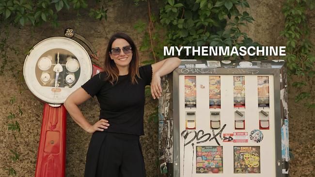© ORF/Neulandfilm Lisa Gadenstätter in schwarzer Kleidung lehnt neben einem Automaten und einer historischen Waage vor einer begrünten Mauer, darüber steht „Mythenmaschine“. Hier bekommt man statt Kaugummis und Spielzeug Mythen. Doch welche spuckt der Automat aus?